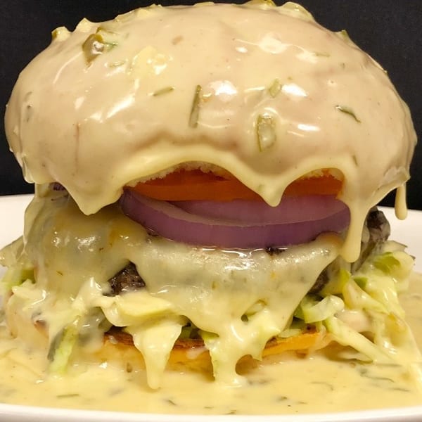 A burger drenched in creamy white sauce, topped with tomato, red onion, and lettuce.