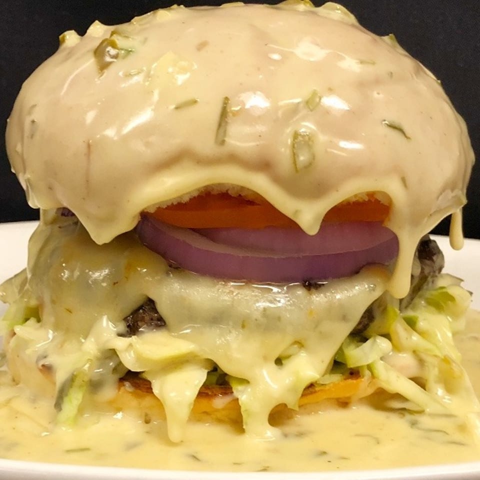 A burger drenched in creamy white sauce, topped with tomato, red onion, and lettuce.
