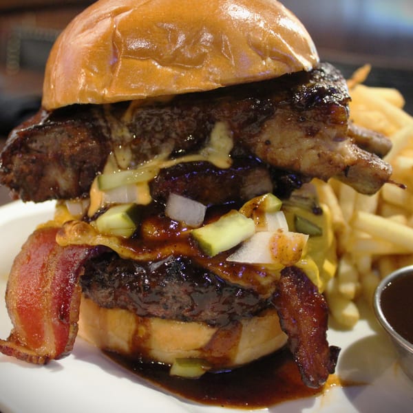 A large burger with bacon, pickles, onions, and barbecue sauce, served with fries and dipping sauce.