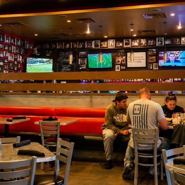 Diners seated at tables and booths in a restaurant with red walls adorned with framed photos and TVs.