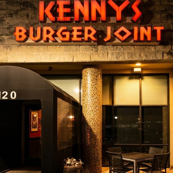 Exterior of Kenny's Burger Joint at night, with glowing red sign and outdoor seating.