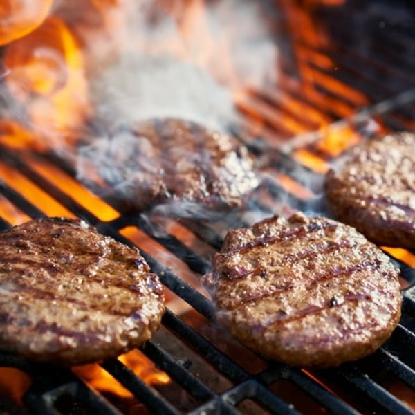 Four sizzling hamburger patties cooking on a fiery grill with smoke rising.