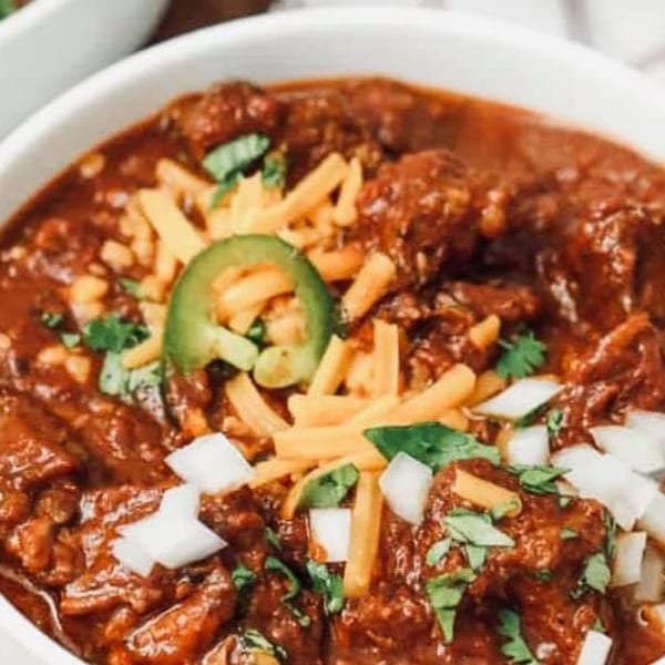 Hearty bowl of chili topped with shredded cheddar, diced onions, cilantro, and a jalapeño slice.