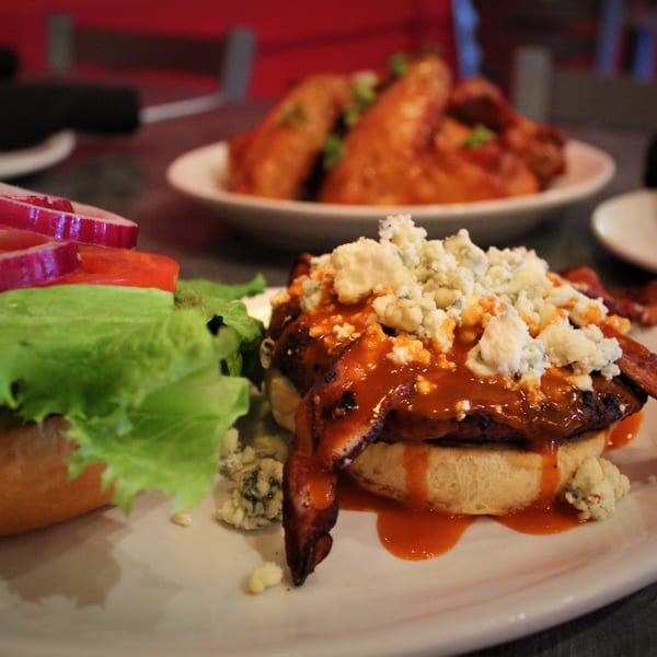 A buffalo chicken sandwich with blue cheese, bacon, lettuce, tomato, and onion, with wings in the background.