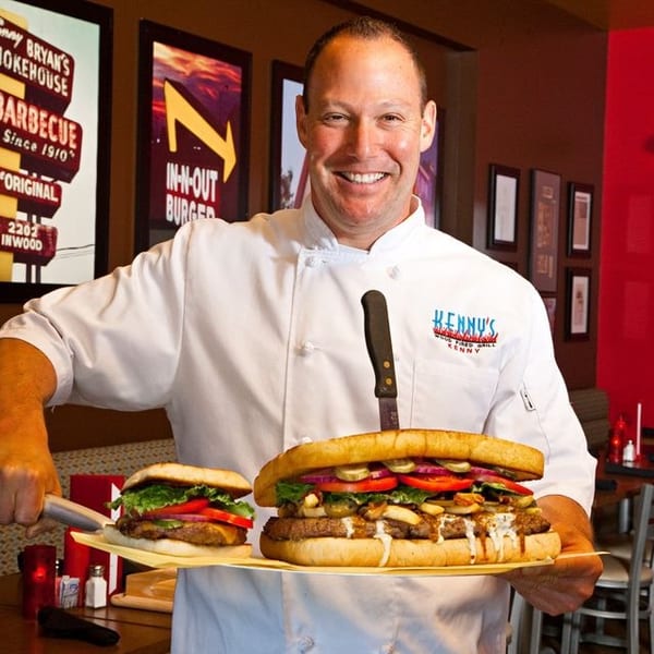 Smiling chef in white uniform holding a giant burger and a regular-sized burger on trays.