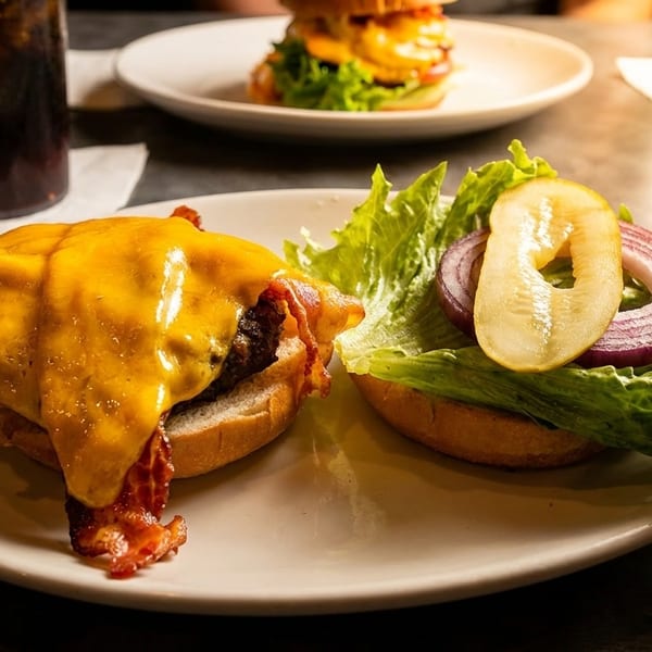 A cheeseburger with bacon, lettuce, red onion, and pickles on a white plate with drinks.